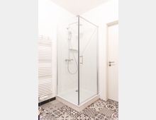 Eckdusche mit Glaswänden und silbernem Rahmen in einem hellen Badezimmer. Der Boden ist mit gemusterten Fliesen gestaltet, und links neben der Dusche befindet sich ein weißer Handtuchheizkörper.