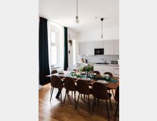 Moderner Essbereich mit einer langen Holztafel, gedeckt mit Geschirr und Dekorationen aus Grünpflanzen. Im Hintergrund befindet sich eine schlichte Einbauküche mit hellgrauen Schränken, einem Herd, einer Mikrowelle und einer Arbeitsplatte vor einer grauen Wand. Ein großes Fenster mit bodenlangen Vorhängen und hängende Glühbirnen beleuchten den Raum.