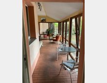 Langer, überdachter Wintergarten mit Ziegelboden, schräger Decke und durchgehender Fensterfront auf der rechten Seite mit Blick in einen kleinen Gartenhof. Im Raum stehen mehrere Sitzgelegenheiten und ein kleiner runder Metalltisch; am Ende befindet sich eine verglaste Tür und weitere Fenster.