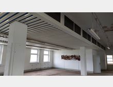 Ein leerer Raum mit weißen Wänden, mehreren Fenstern und einer Decke mit Metallverstrebungen und eingelassenen Gitteröffnungen. Die Wände zeigen Abnutzungsspuren, und eine hängende Neonbeleuchtung sowie ein beschädigtes Wandobjekt sind sichtbar.