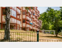 Mehrstöckiges Wohngebäude mit orangefarbenen Balkonstrukturen, umgeben von einer Grünfläche mit Bäumen und Rasen. Im Vordergrund ist ein Birkenbaum sowie ein Zaun erkennbar, im Hintergrund Wäscheleinen auf der Grünfläche.