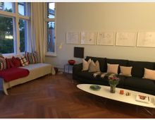 Wohnzimmer mit Parkettboden, einer großen dunklen Couch mit hellen Kissen und einem ovalen weißen Couchtisch. Links befindet sich eine Tagesliege mit bunten Kissen vor hohen Fenstern mit weißen Vorhängen. An der Wand hängen mehrere gerahmte Zeichnungen.