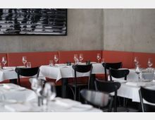 Ein moderner Restaurantinnenraum mit gedeckten Tischen, weißen Tischdecken, schwarzen Stühlen und einer roten Polsterbank entlang der Betonwand. An der Wand hängt ein abstraktes Kunstwerk in Schwarz-Weiß.