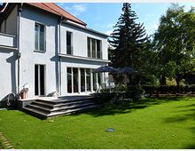 Moderne zweigeschossige Villa mit weißer Fassade, großen bodentiefen Fenstern und einem roten Satteldach. Im Vordergrund ein gepflegter Garten mit Rasen, Terrassenstufen und einem Sonnenschirm, umgeben von Bäumen und Sträuchern.