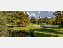 Parklandschaft mit grünen Wiesen, großen Bäumen und einem schmalen Fluss im Vordergrund; im Hintergrund ist der Turm einer Kirche zu sehen. Moderner Sitzbereich mit Betonflächen und Sitzgelegenheiten im mittleren Teil des Parks.