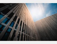 Moderne Gebäudefassade aus vertikalen Holzlatten mit großen Fenstern, die blauen Himmel und Sonnenlicht reflektieren; das Bild zeigt die Architektur aus einer niedrigen Perspektive mit Blick nach oben.