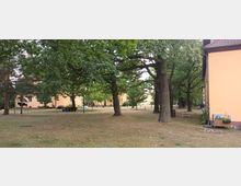 Rasenfläche mit großen Bäumen, im Hintergrund mehrere gelb verputzte Gebäude mit roten Dächern. Rechts im Bild ist die Außenwand eines Gebäudes sichtbar; Holzbänke und ein kleiner Spielbereich befinden sich unter den Bäumen.