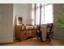 Heller Wohnraum mit Holzfußboden, großem Fenster mit transparenten Vorhängen und einem niedrigen Holzsideboard mit Dekoration. Daneben steht ein dunkler Stuhl mit Kissen und übergehängtem Tuch; eine Zimmerpflanze ragt am rechten Bildrand ins Bild.