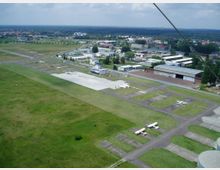 Kleiner Flughafen mit mehreren asphaltierten Start- und Landebahnen, parkenden Kleinflugzeugen sowie einem Kontrollturm und angrenzenden Hangars; umgeben von grünen Wiesen und einzelnen Gebäuden im Hintergrund.