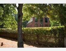 Backstein-Haus mit großen, grün gerahmten Fenstern hinter einer Hecke und einem Holzzaun, umgeben von Bäumen und sommerlicher Vegetation. Im Vordergrund steht ein Baum, der teilweise das Gebäude überschattet.