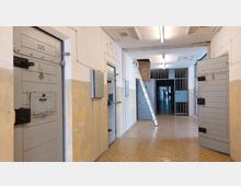 Ein Gefängnisflur mit massiven grauen Zellentüren, die nummeriert und mit Schlössern sowie kleinen Sichtklappen versehen sind. Der Raum ist schlicht gestaltet, mit abblätternder Wandfarbe, einem gemusterten Bodenbelag und einer Leiter an der Wand; im Hintergrund sind Gittertüren und ein weiterer Flur sichtbar.