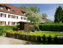 Wohnanlage mit weiß gestrichenem Gebäude, rotem Satteldach und braunen Fensterläden, umgeben von einem Garten mit Holzlaube, blühendem Baum und Rasenflächen, eingefasst durch einen Holzzaun und Hecken.