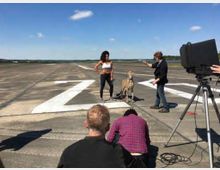 Eine Szene auf einer Landebahn mit einem Fotoshooting: Eine Frau steht neben einem Geparden, während mehrere Personen im Vordergrund das Shooting vorbereiten. Ein Fotograf und eine Kamera auf einem Stativ sind sichtbar, der Himmel ist klar und blau.