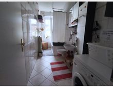 Narrow, tiled bathroom with a bathtub and white shower curtain at the far end beneath a window. A wall-mounted toilet and pedestal sink line the right side, with a washing machine in the foreground and striped rugs on the tiled floor.