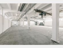 Großer, leerer Industriebau mit weißen Wänden, sichtbaren Stahlträgern und Betonboden. Die Halle hat eine hohe Decke mit einem Schienensystem für Hebevorrichtungen und große Fenster entlang der Seitenwände, die viel Tageslicht einlassen.