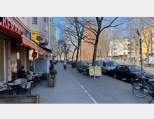 Straßenszene in einer Stadt mit Fußgängerweg, auf dem links ein Café mit Tischen und Stühlen vor der Fassade eines Gebäudes steht. Rechts Bäume, geparkte Autos und Fahrräder entlang der Straße; im Hintergrund Mehrfamilienhäuser und ein rotes Backsteingebäude sichtbar.