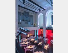 Innenansicht eines modern umgestalteten Kirchengebäudes mit hohen Decken, großen Rundbögen und einer Orgel an einer Wand. Der Raum ist in farbigem Licht beleuchtet und mit weißen Tischen, Stühlen sowie Dekorationen für eine Veranstaltung ausgestattet.