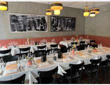 Ein modernes Restaurant mit gedeckten Tischen, weißen Tischdecken und schwarzen Stühlen. Der Raum hat eine graue Betonwand mit abstrakter Kunst und orangefarbenen Sitzpolstern entlang der Wand.