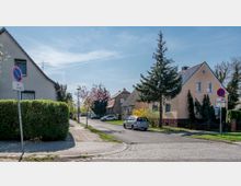 Ruhige Wohnstraße mit Einfamilienhäusern und Gärten, flankiert von Gehwegen und vereinzelten Bäumen. Im Vordergrund stehen Verkehrsschilder mit Halteverbotshinweisen, und parkende Autos sind entlang der Straße zu sehen.