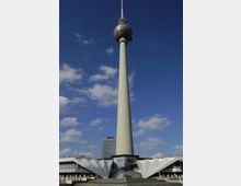 Der Berliner Fernsehturm bei Tag, mit klarem Himmel und wenigen Wolken im Hintergrund. Im Vordergrund befindet sich das flache, weiße Gebäude des Eingangsbereichs mit modernen, skulpturalen Elementen, und im Hintergrund ist das Park-Inn-Hotel zu sehen.