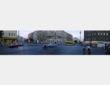 Straßenszene in einer Stadt mit einem mehrspurigen Verkehrsknotenpunkt, durchzogen von Straßenbahnschienen. Umgeben von mehrstöckigen Wohn- und Geschäftshäusern, darunter ein gelbes Gebäude mit einer Apotheke im Erdgeschoss. Im Hintergrund ist der Berliner Fernsehturm sichtbar.