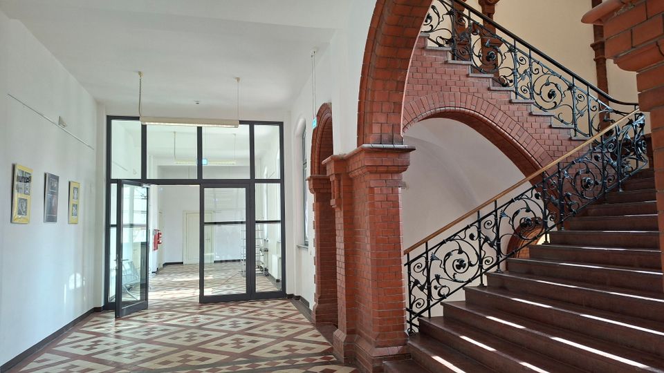 Heller Innenflur eines Gebäudes mit gemustertem Fliesenboden, weißen Wänden und einer Glas-Doppeltür zu einem weiteren Raum. Rechts führt ein breites Treppenhaus mit roten Ziegelbögen und dekorativem schmiedeeisernem Geländer nach oben.