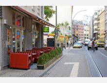 Straßenszene in einer Stadt mit einem Diner im Vordergrund, dessen rote Sitzbänke im Außenbereich stehen, neben einer mit Pflanzen gefüllten Holzkiste. Im Hintergrund sind eine belebte Straße mit Fußgängern, Autos, Fahrradfahrern und moderne, teils bunte Gebäude zu sehen.