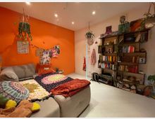 Wohnzimmer mit einer orangefarbenen Wand, grauem Ecksofa, dekorativen Kissen und bunten Wandbildern. Auf der rechten Seite ein großes Bücherregal, Pflanzen, eine Wanduhr und helle Bodenplatten.