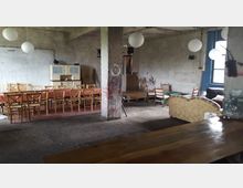 Ein großer, rustikaler Raum mit unverputzten Wänden, einem zentralen Betonpfeiler und einfacher Einrichtung. Im Vordergrund stehen Tische mit Stühlen, hinten befinden sich Vintage-Möbel und eine gemütliche Sitzecke mit Sofas.