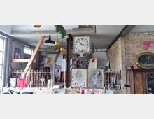 Ein Atelier mit einer offenen, industriellen Atmosphäre, geprägt von unverputzten Ziegelwänden und einer Vielzahl an Kunstwerken wie Gemälden und Zeichnungen, die den Raum dekorieren. Eine große Uhr ist ein zentraler Blickfang, und Regale sowie Möbelstücke ergänzen die kreative Umgebung.