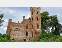 Verfallenes Backsteingebäude mit einem hohen Turm und gotischen Fenstern, umgeben von Bäumen und dichter Vegetation; die Fassade zeigt Spuren von Verwitterung und Bewuchs.