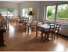 Heller Speiseraum mit Holzboden, ausgestattet mit mehreren Holztischen und -stühlen, die mit weißen Tischdecken gedeckt sind. Große Fenster auf zwei Seiten bieten Blick auf eine grüne Außenanlage und eine hölzerne Terrasse.