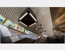 Blick auf den oberen Bereich einer U-Bahn-Station mit breiten, metallverkleideten Rolltreppen. Die Decke ist mit weißen Holzpaneelen und rechteckigen, beleuchteten Leuchten ausgestattet; im Hintergrund sind bunte Wohngebäude und gelbe Stützen sichtbar.