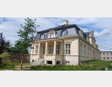 Historisches Gebäude im klassizistischen Stil mit einem Mansardendach, großen Fenstern und einer überdachten Veranda, die von Säulen getragen wird. Das Gebäude steht in einer Grünfläche, umgeben von einer niedrigen Mauer und angrenzenden modernen Strukturen.