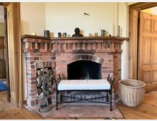 Gemütlicher Innenraum mit einem gemauerten Kamin aus roten Ziegelsteinen. Der Kamin besitzt eine Nische für Holzlagerung, dekorative Gegenstände auf dem Kaminsims, ein hell gepolstertes Sitzmöbel davor und einen geflochtenen Korb daneben.