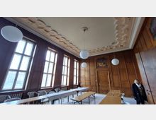 Ein Raum mit holzgetäfelten Wänden, großen Sprossenfenstern und einer dekorativen Decke mit geometrischen Mustern. Es stehen Holz- und Kunststofftische sowie Stühle, und von der Decke hängen runde, weiße Pendelleuchten.