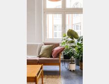 Ein helles Wohnzimmer mit einem Sofa aus Holzrahmen und beigen Polstern, geschmückt mit grünen und rosa Kissen. Im Vordergrund ein Holztisch, daneben ein Teppich, und im Hintergrund mehrere grüne Zimmerpflanzen vor einem großen Fenster mit Blick auf eine helle Fassade.