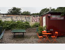 Ein Außenbereich mit Kiesboden, einer Tischtennisplatte, einem roten Containergebäude mit Werbebanner und einem kleinen Set aus orangenen Gartenmöbeln. Im Hintergrund eine mit Graffiti bedeckte Mauer und üppige grüne Sträucher.