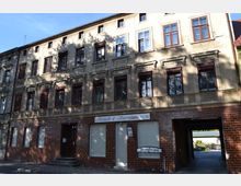 Altbau-Wohngebäude mit drei Etagen, verzierten Fassadenelementen und rötlich-brauner Klinkerfassade im Erdgeschoss. Im Erdgeschoss befindet sich ein Ladengeschäft mit der Aufschrift "Fleisch & Wurstwaren" und ein Durchgang zu einem Innenhof.