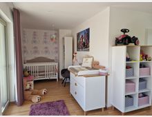 Helles Kinderzimmer mit Holzfußboden, einem Gitterbett, einem Wickeltisch und Regalen mit farbigen Aufbewahrungsboxen. An der Wand befinden sich verspielte Tapeten mit Elefantendesign, und ein großes Stofftier liegt auf dem Boden.