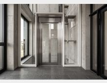 Moderner Innenraum mit einem Aufzug in Edelstahloptik, eingebettet in eine Betonwand. Große Fenster mit schwarzen Rahmen sowie Glas- und Metallelemente umgeben das Aufzugsmodul, der Boden ist mit dunklen Fliesen ausgelegt.