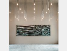 Minimalistischer Galerieraum mit Betonboden, einer großformatigen abstrakten Kunstmalerei an der beige gestrichenen Wand und zahlreichen hängenden Pendelleuchten mit warmem Licht, die von der Decke herunterreichen.