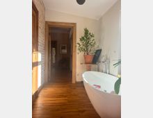 Ein Badezimmer mit freistehender, weißer Badewanne, Holzboden und einer Ziegelwand auf der linken Seite. Im Hintergrund führt ein Türrahmen aus Holz in einen Flur, und ein großer grüner Blumentopf steht auf einem kleinen Holztisch neben der Wanne.