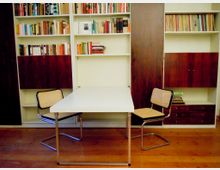 Arbeits- oder Leseecke mit weißem Tisch und zwei Freischwinger-Stühlen vor einer wandfüllenden Einbau-Bücherwand. Die Regale sind mit Büchern gefüllt und haben dunkelholzfarbene Schranktüren und Schubladen über einem Holzboden.