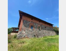 Stein- und Ziegelgebäude mit einer Fassade aus Naturstein im unteren und rotem Ziegel im oberen Bereich, mit mehreren kleinen Fenstern; das Gebäude steht auf einer Wiese bei sonnigem Wetter mit blauem Himmel. Links ist eine überdachte Veranda zu erkennen.
