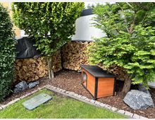 Ein gepflegter Gartenbereich mit einem kleinen, hölzernen Tierhaus rechts im Bild, umgeben von Bäumen, Steinbrocken und gestapeltem Brennholz an einer weißen Wand. Im Vordergrund verläuft ein grüner Rasen, der von einer gepflasterten Kante begrenzt ist.