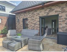 Gepflasterte Terrasse an einem Haus mit Klinkerfassade und grauem Ziegeldach, ausgestattet mit einer Loungegarnitur aus grauem Rattan und Couchtisch. Rechts führt eine große, geöffnete Schiebetür in den Wohnbereich; links ist ein angrenzender Holzschuppen mit Wandlampe zu sehen.