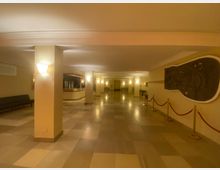 Heller Eingangsbereich mit beige gefliesten Böden und einer Rezeption im Hintergrund. An den Wänden befinden sich Wandlampen und ein großes Relief, das von roten Absperrkordeln abgegrenzt ist.