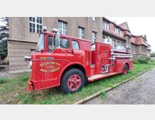 Rotes historisches Feuerwehrauto mit der Aufschrift „Beckemeyer F.P.D Wade Township“, geparkt auf einer Grünfläche vor einem beige-braunen Gebäude mit roten Ziegeldächern und Sprossenfenstern.