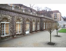 Ein historisches Gebäude mit großen, bogenförmigen Fenstern, die von kletternden Pflanzen umrahmt sind. Im Vordergrund befinden sich ein gepflasterter Hof mit kleinen, unbelaubten Bäumen und grüne Rasenflächen, während im Hintergrund Wohngebäude sichtbar sind.